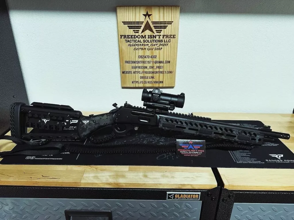 A tactical firearm with a mounted scope rests on a mat atop a workbench, with a sign in the background for "Freedom Isn't Free Tactical Solutions LLC." The workbench has metal drawers at the bottom, and various tactical accessories are nearby.