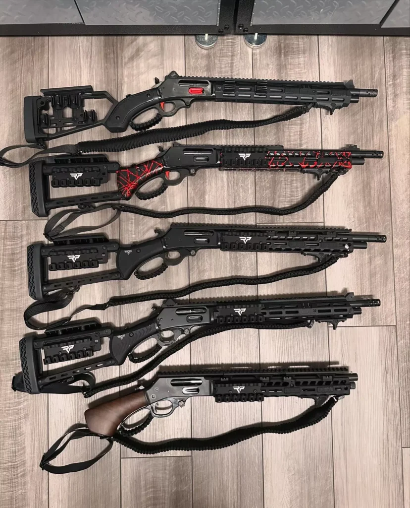 Six customized lever-action rifles with tactical features, such as upgraded stocks, rails, and red accents, are lined up on a wooden floor. The rifles are arranged next to each other with their barrels facing the same direction.