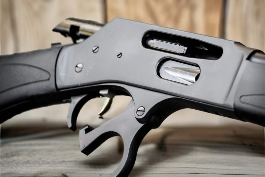 Close-up image of a black lever-action rifle with a partially open action, showcasing its intricate metal components and trigger mechanism against a blurred wooden background.